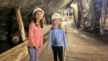 Two kids are on a family trip in the Northeast in the state of Pennsylvania. Two girls in hard hats stand underground in a lit coal mine with rock walls and wooden flooring in Pennsylvania.