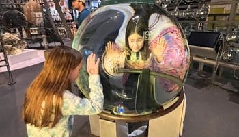 Girls on a family trip in Upstate NY are having fun at a museum. Two girls gaze into a giant glass ball at one another in a museum exhibit at the Corning Museum of Glass in Upstate NY.