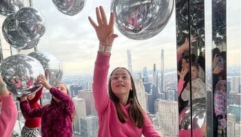 Kids on a New York City small group tour visit the Summit One iconic site. A girl reaching for a floating silver ball with New York City skyline visible in the windows behind her, with other kids holding and reaching for silver balls nearby.