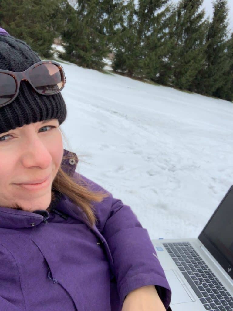A woman, Amanda Cave Jackson, working with laptop in a snowy yard with winter coat and hat on.