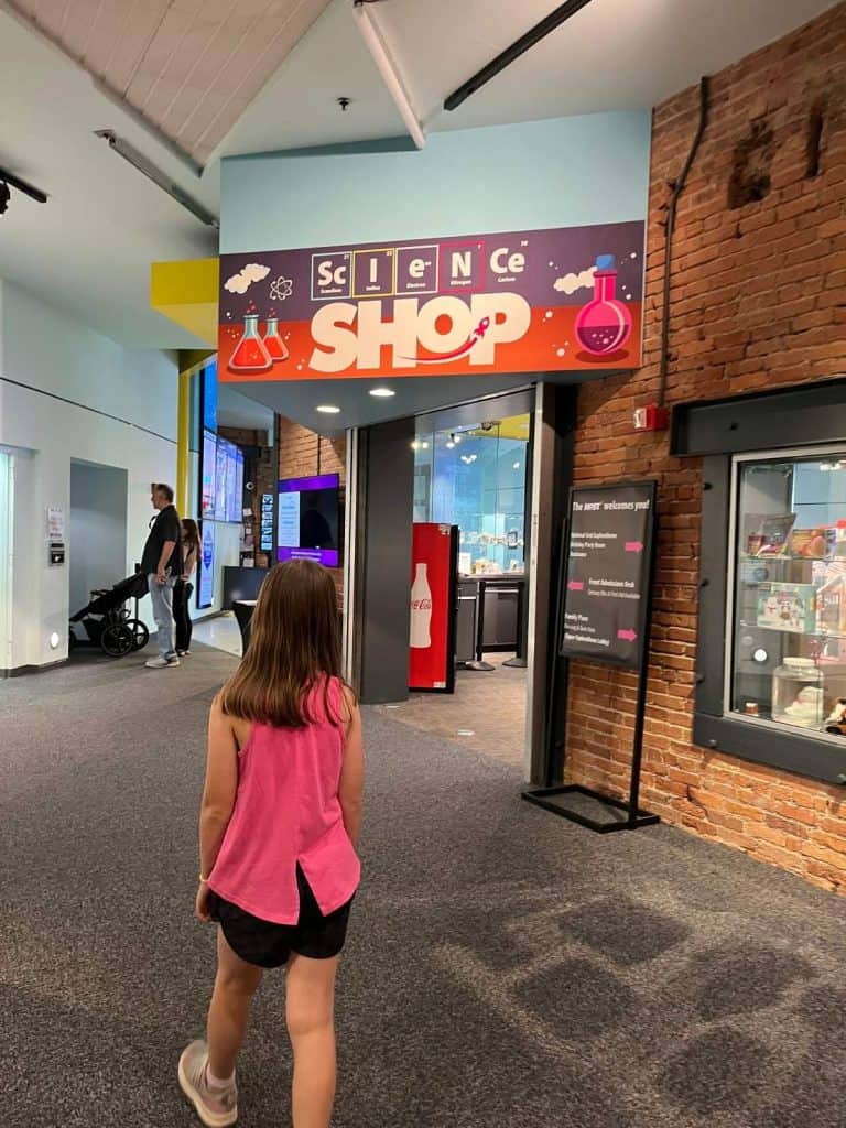 A girl walks toward an alcove with brick exterior that has a Science Shop sign with chemical bottle graphics.