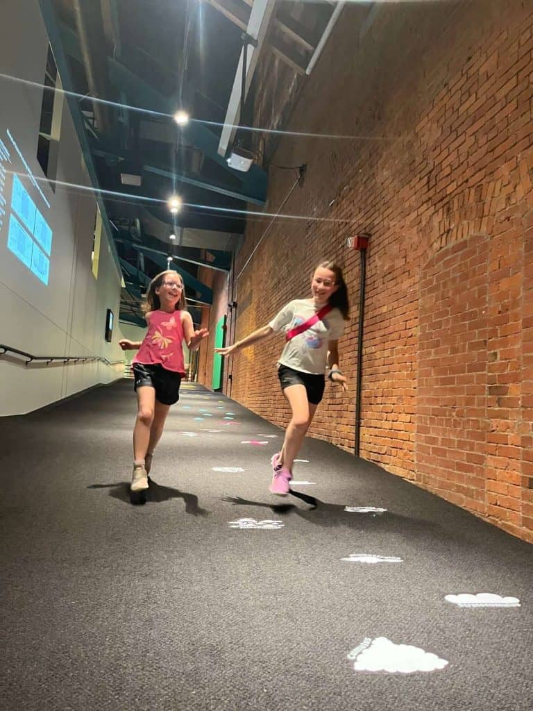 Two girls run down a brick hallway with carpet and decals in the Syracuse Science Museum.