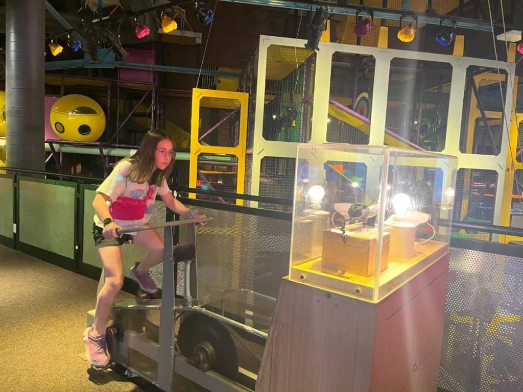 A girl rides an indoor bike that lights up a lightbulb in a clear case in front of the bike.