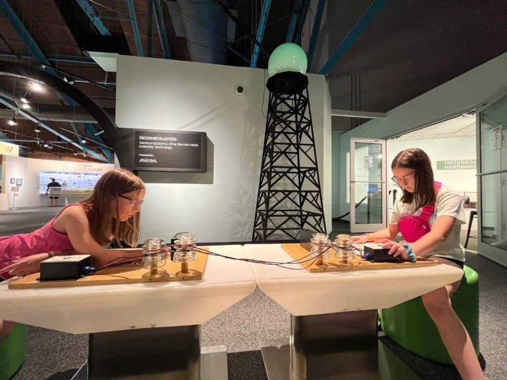 Two girls sit on either end of a table at a Morse Code station in front of a replica communications tower.