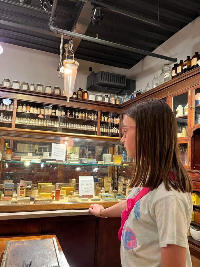A girl looks at a clear case with bottles and jars showing medicines and lotions, with wooden displays along the walls of glass jars with tonics and potions.