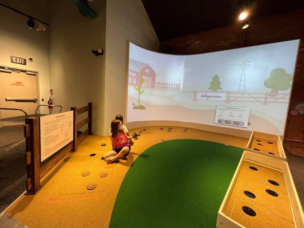Two girls look at a projection screen showing a farm, and plant and pull circular plants from brown carpet made to look like soil near green grass carpet.