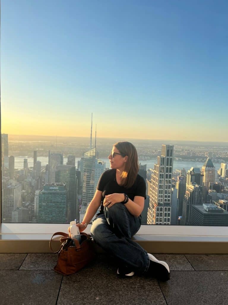 A woman sitting on a rooftop looking to the left with New York City skyline in the background at dusk with blue sky and yellow horizon.