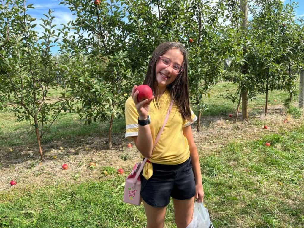 A girl in a yellow shirt smiles while holding an apple in an orchard in New York.