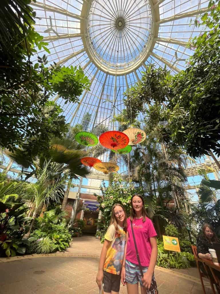 Two girls inside the Buffalo Botanical Garden with lush greenery, beneath a vaulted glass ceiling with colorful umbrellas overhead.