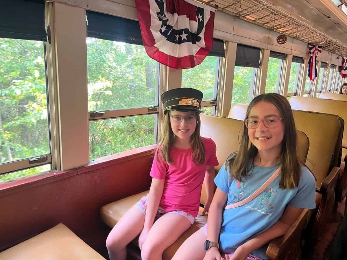 Two girls sit on a Cooperstown train, one wearing a conductor hat.