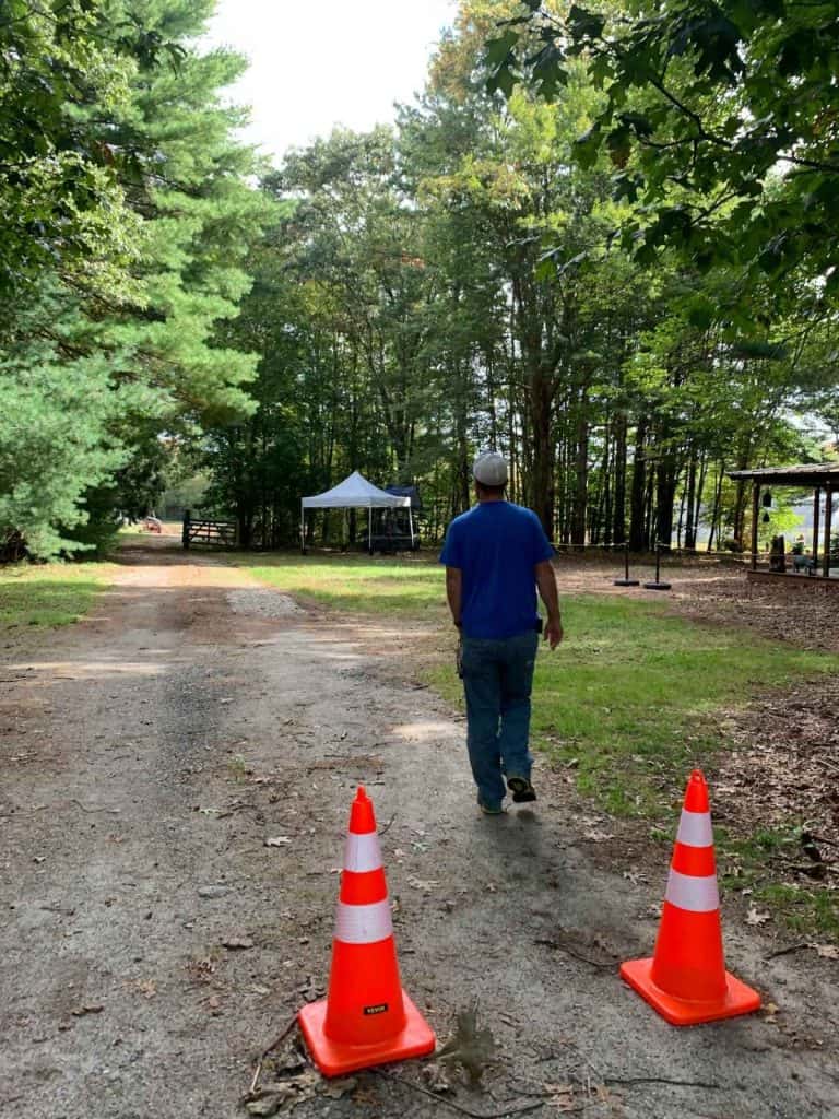 A man walks down a dirt/gravel path past two cones in a wooded area towards some tents and cranberry fields in the far distance.