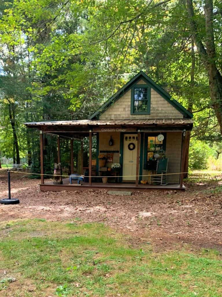 A small cottage called Cranberry Cottage sits in a wooded area near a cranberry bog tours area with a front porch, rocking chairs, and rustic decorations.
