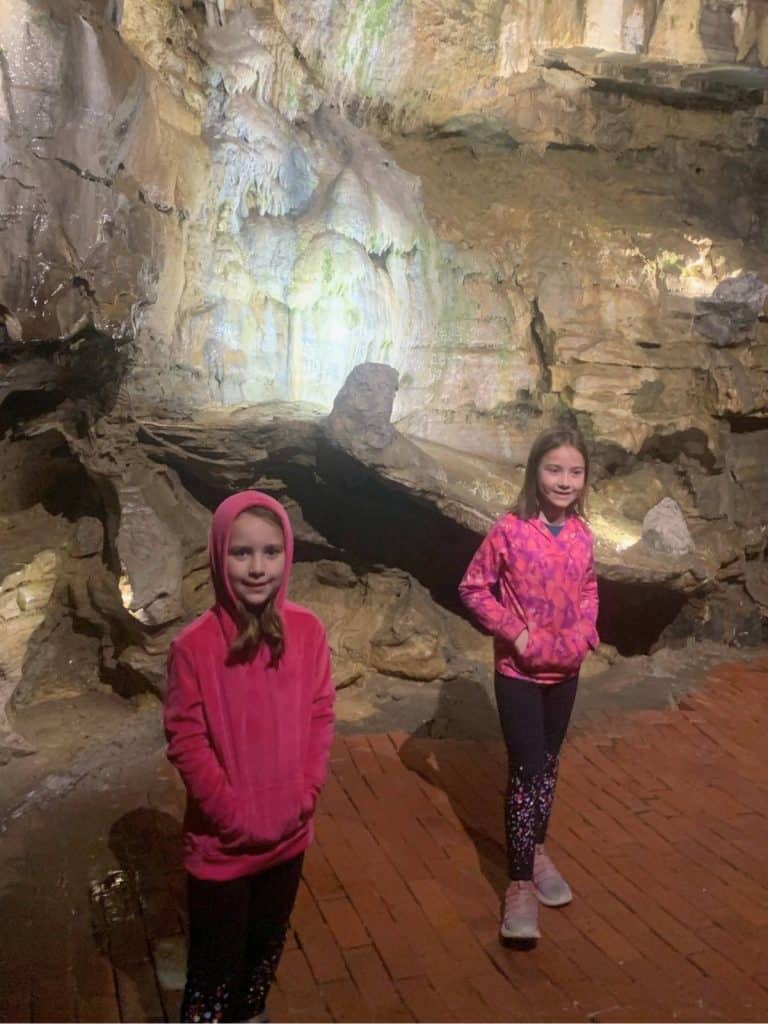 Two girls in pink sweatshirts standing in front of a backlit rock formation in an underground cave on a family hike at Howe Caverns.