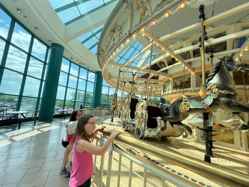 Two girls look at a carousel enclosed in a glass atrium within a mall on a bright sunny day.