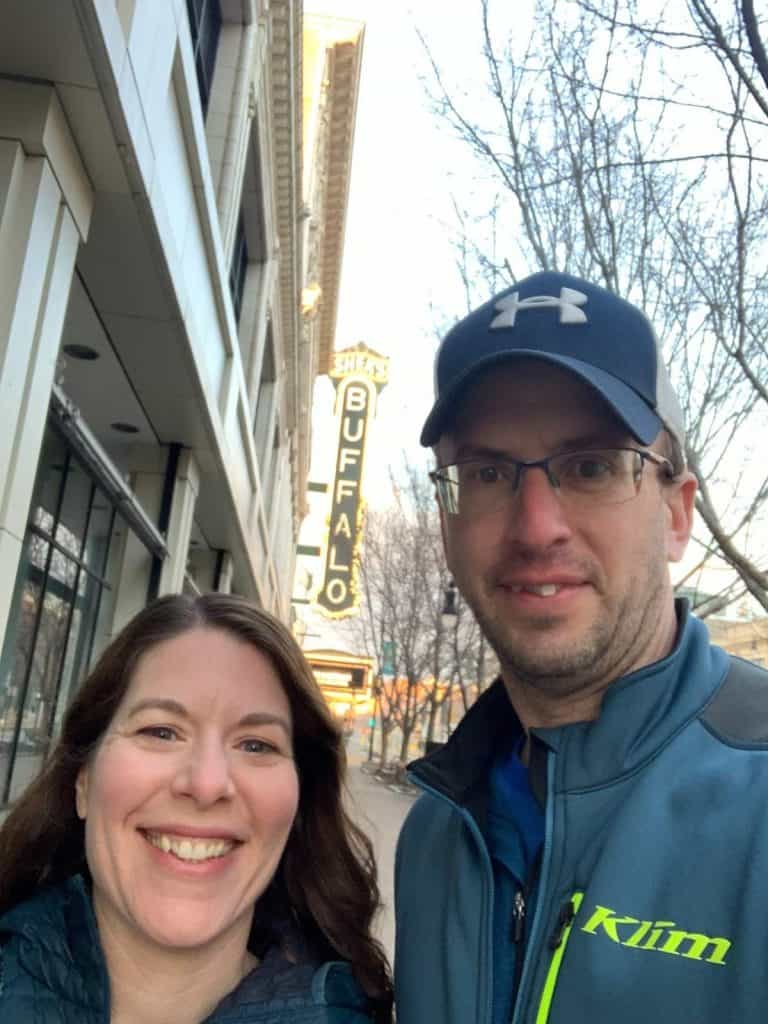 A man and woman standing on a tree-lined sidewalk area outside an illuminated historic sign that says Shea's Buffalo in the theatre district in Upstate NY. 