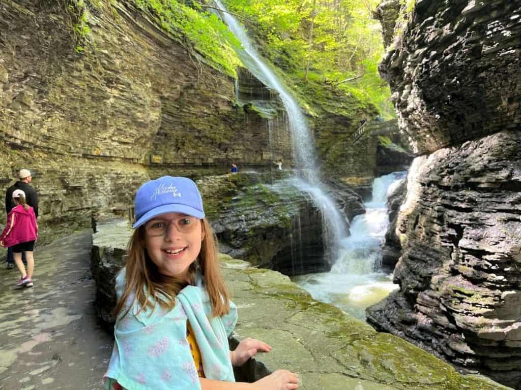 A girl in a hat and glasses standing in front of a waterfall and a stone trail at Watkins Glen State Park.