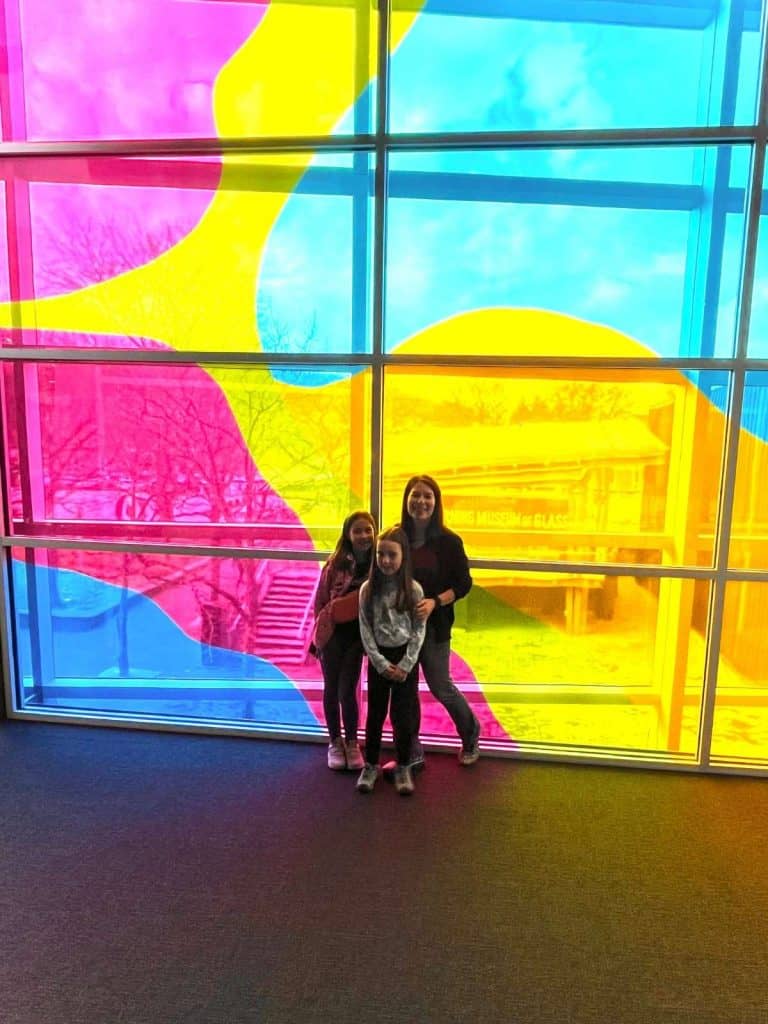 A mom and two kids standing in front of a window with large blue, pink, and yellow swirls at the Corning Museum of Glass in upstate NY.