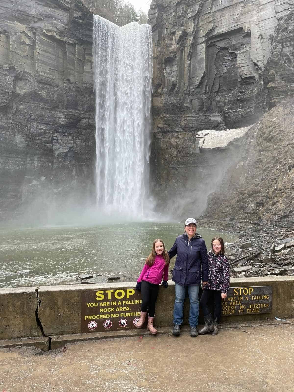 A woman and two girls in front of Taughannock Falls State Park large waterfall attraction surrounded by rock walls.