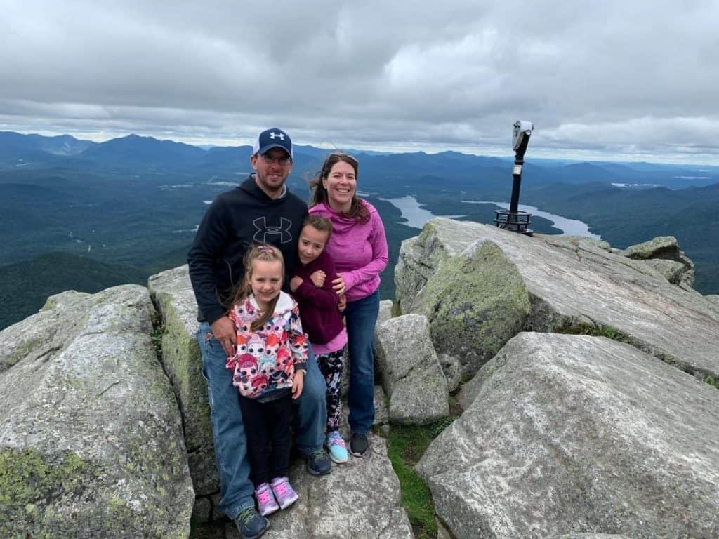 A family of 4 standing on large boulders at the top of Whiteface Mountain in the Adirondacks of NY, huddled together with the breeze blowing their hair.