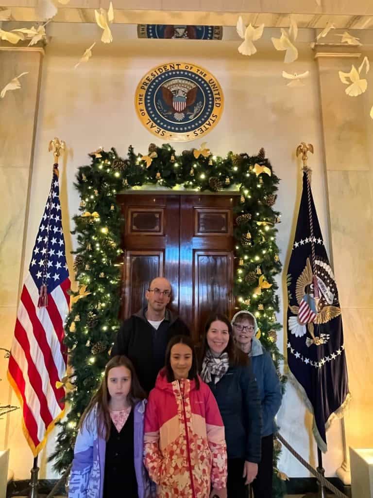 A family of 5 in front of a dark wood door and next to American flags, below the White House Presidential Seal in a hallway surrounded by white marble tiles.