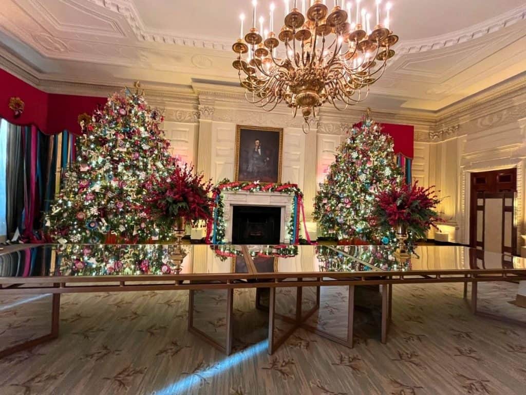 The State Dining Room viewed on a White House Tour is decorated with two large colorful Christmas trees, an ornate fireplace with surrounding white paneling, and a large, mirrored table.