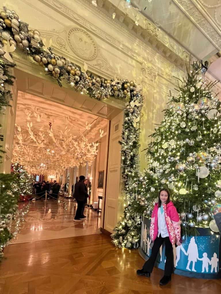 A smiling girl in a pink jacket stands in front of a 12-foot decorated Christmas tree and a hallway with a ceiling decorated with doves on a White House tour.