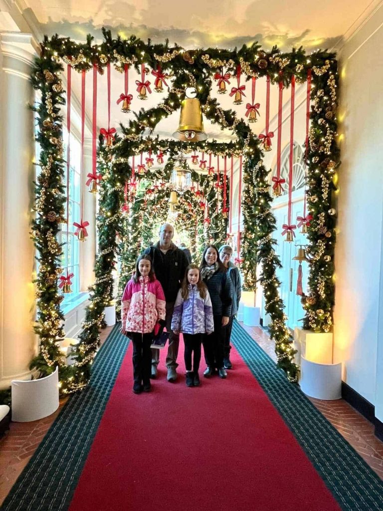 A family of 5 smiles in the hallway of the White House with red and green carpet, windows, and holiday evergreen garland with gold bulbs and red string with gold bells hanging from the ceiling.