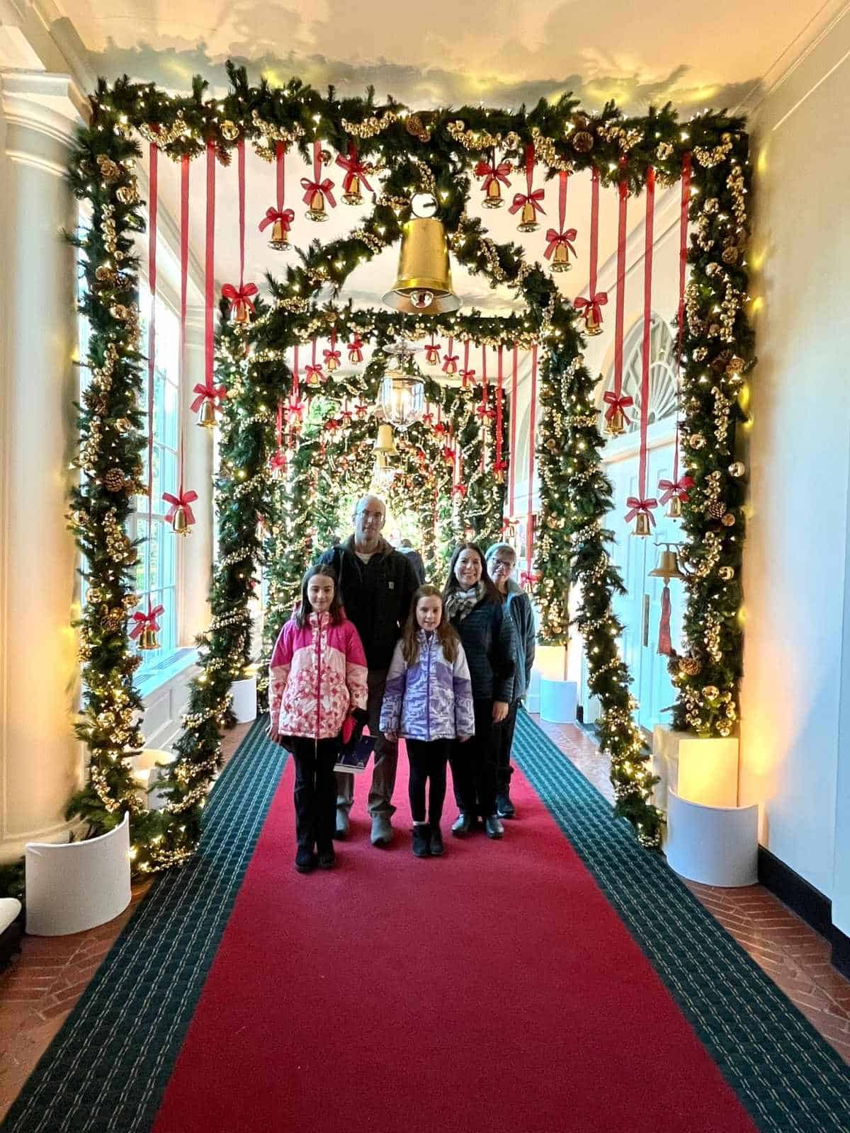 A family of 5 smiles in the hallway of the White House with red and green carpet, windows, and holiday evergreen garland with gold bulbs and red string with gold bells hanging from the ceiling.