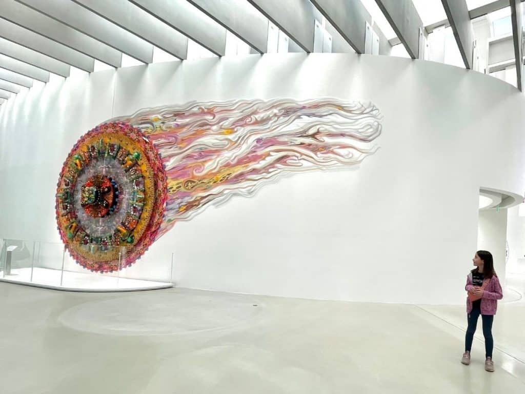 A young girl stands in a bright, white modern gallery, looking up at a massive, vibrant glass sculpture that resembles a colorful comet or sun trailing white wisps across a curved wall.