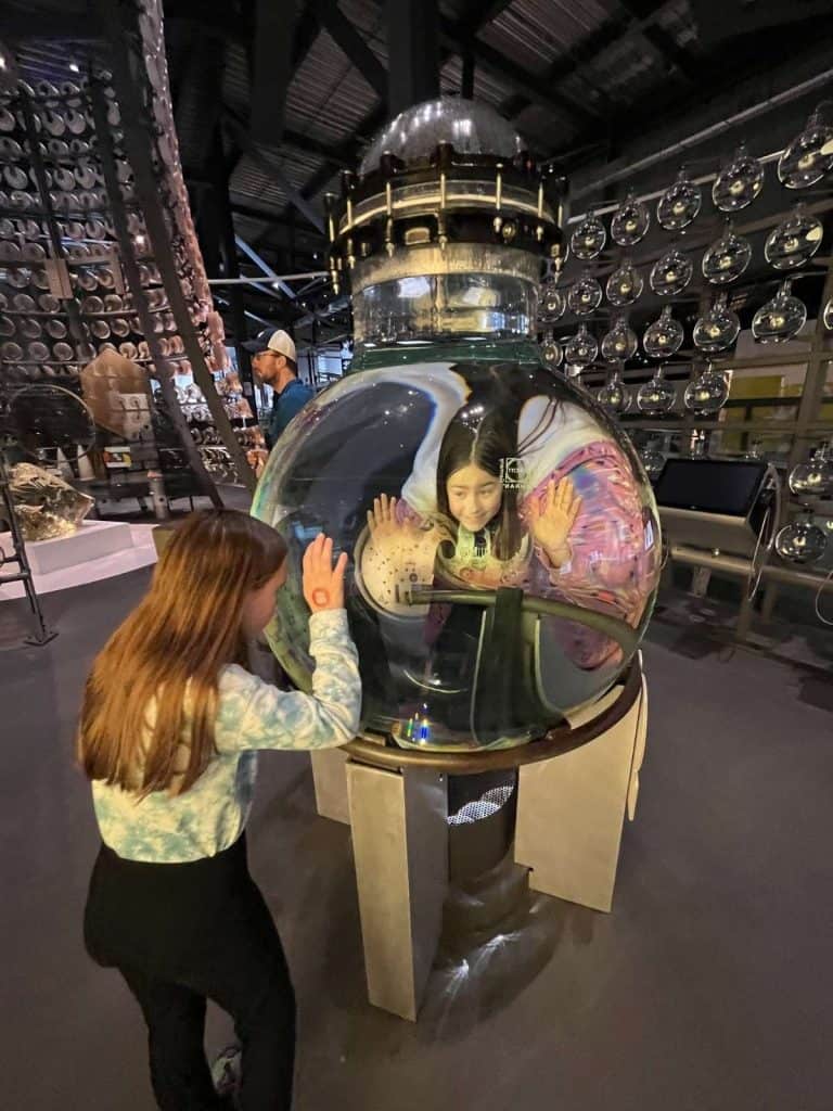 Two girls looking through a giant glass sphere, with a wall grid of smaller glass spheres suspended behind them.