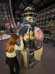 Two children interact with a large, hands-on glass globe exhibit, peering through the glass while touching its surface. In the background, dozens of smaller glass orbs are suspended from the ceiling in a precise geometric pattern.