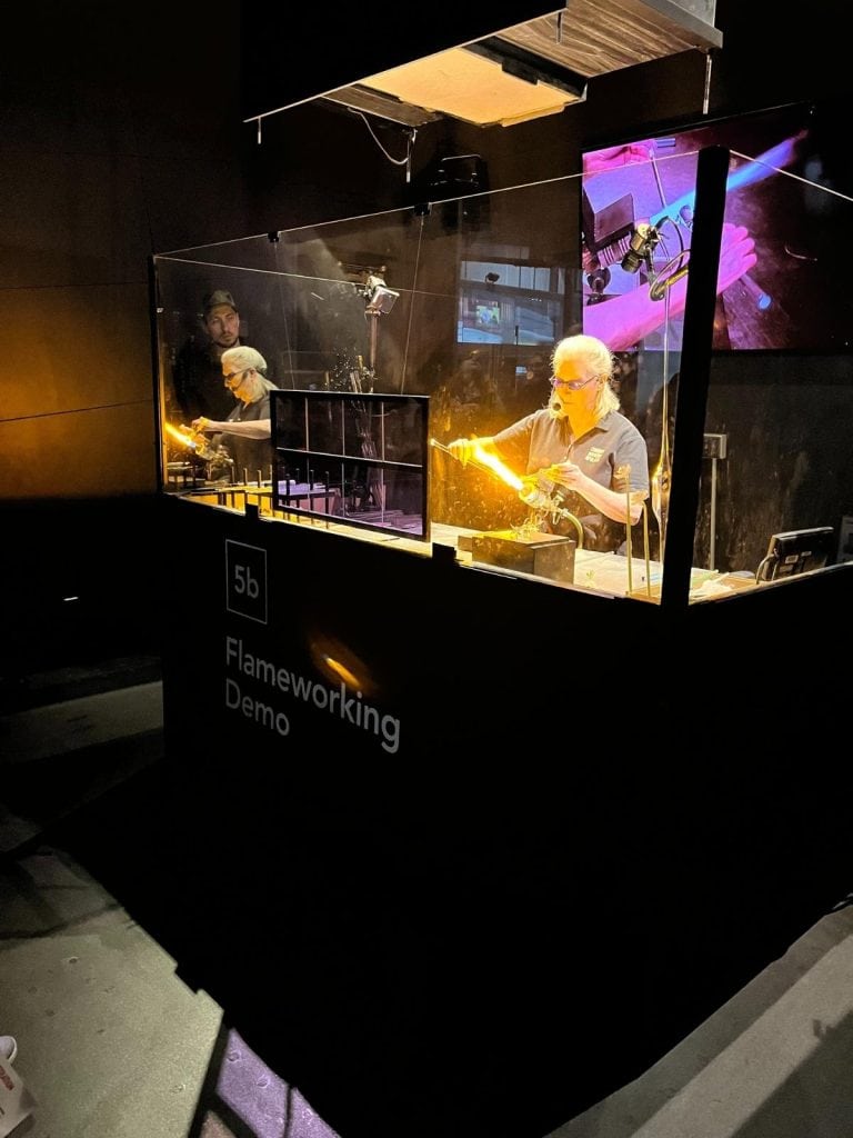 A glassblowing artist shaping molten glass behind safety glass during a live flameworking demo with flame and glass rod.