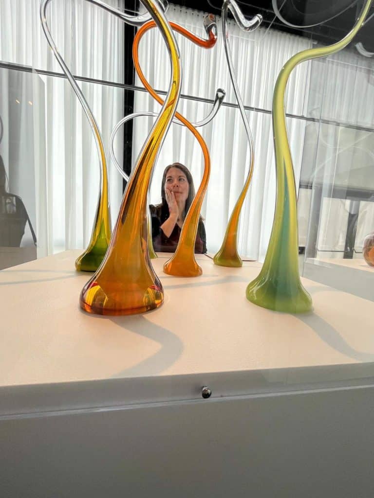 Woman stands next to winding whimsical glass sculptures in bright green and orange.