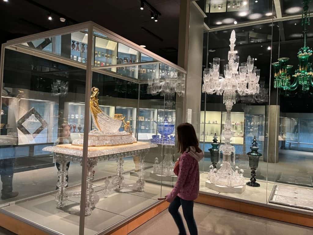 Child observes detailed glass chandeliers and historical glass art lit up in the dark museum’s main gallery.