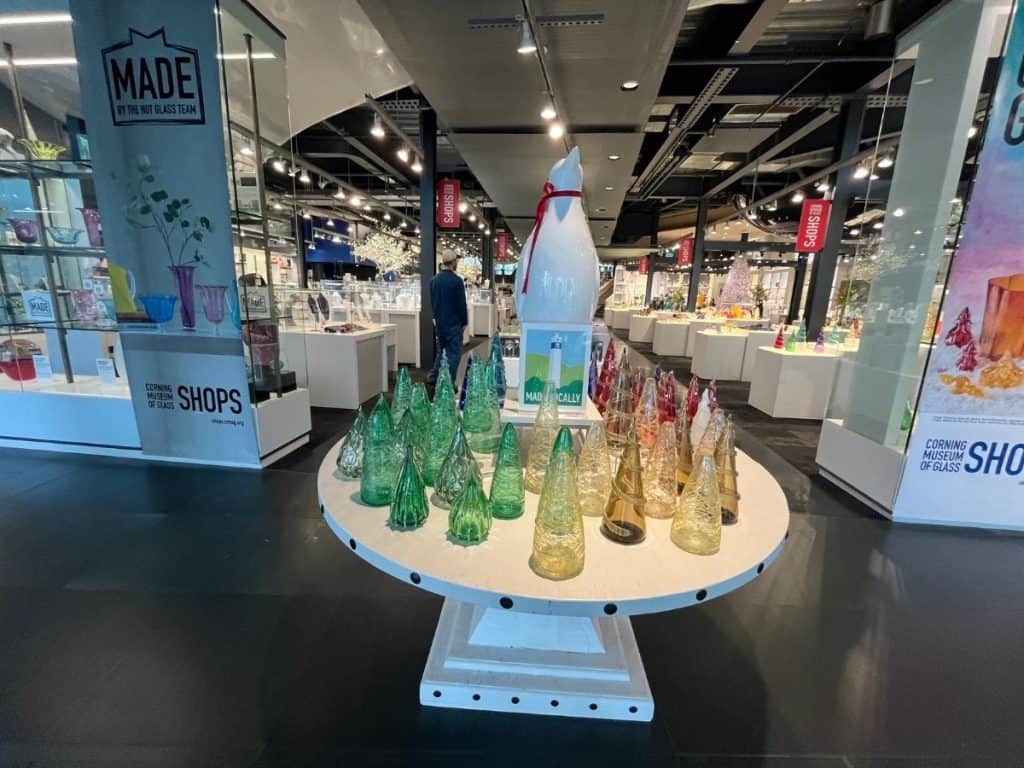 A display of brightly colored glass trees surrounding a giant penguin centerpiece, situated in a modern museum gallery with a large, well-lit gift shop in the background.