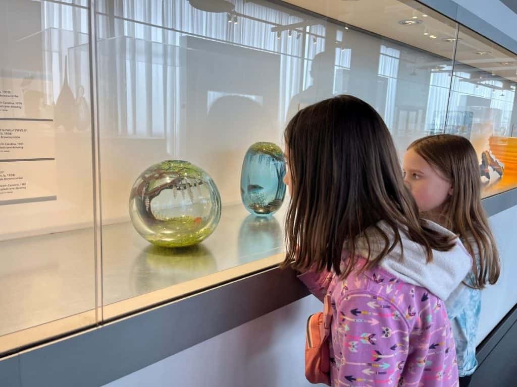 Two girls closely examine glass orbs and delicate sculptures in a well-lit display case.