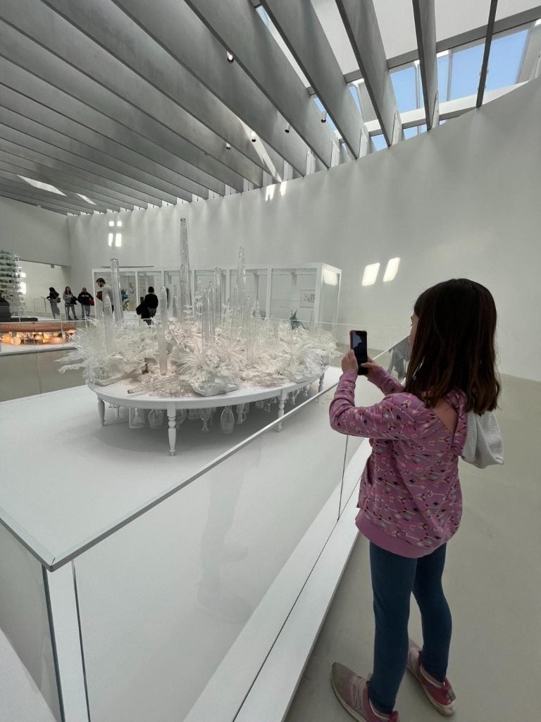 A young girl photographs an elaborate white glass sculpture in a sunlit gallery.