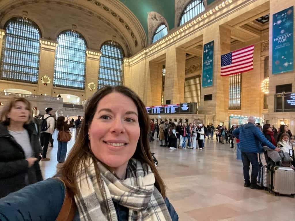 A selfie of a smiling woman with brown hair and a plaid scarf in the Main Concourse of Grand Central Terminal. The background features the terminal's iconic high arched windows, the green celestial ceiling, and a large American flag. Banners for a 'Holiday Fair' are visible, along with crowds of travelers and wreaths on the walls.