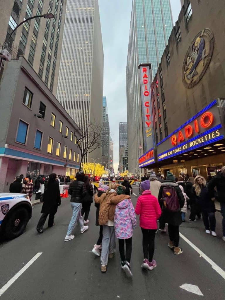 A group of school aged kids walks down the road toward Radio City Music Hall, with other pedestrians nearby.