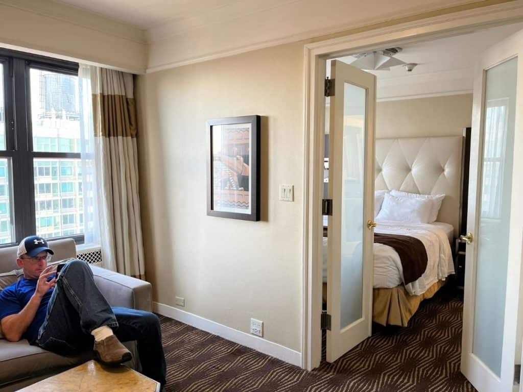 A man sitting in a hotel suite living area with double doors leading to a bed with a tufted headboard and white crisp sheets.