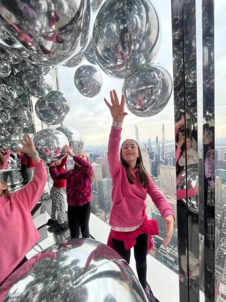 A girl in a pink sweater reaches for a silver floating ball at Summit One with the NYC skyline behind her.