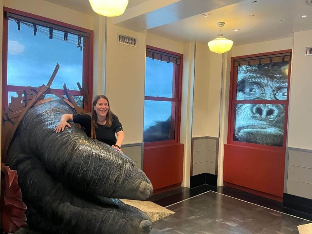 A woman stands in the fingers of a King Kong Hand while he looks in the window at the Empire State Building.