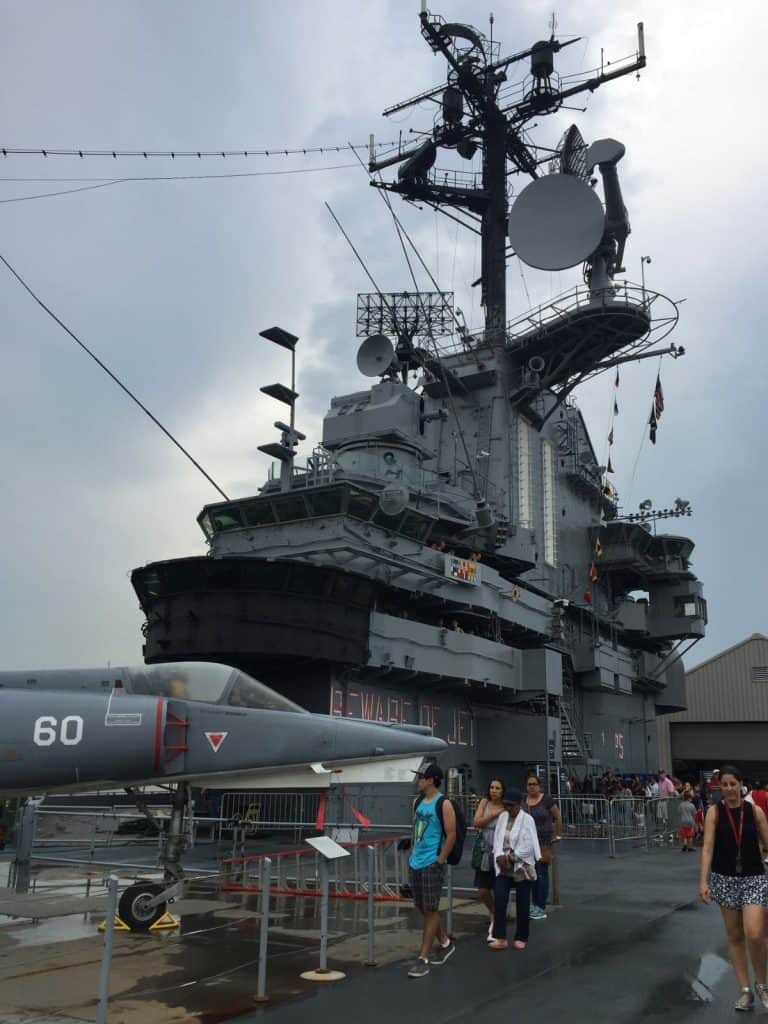 The radar equipment of the USS Intrepid aircraft carrier, with a vintage fighter jet parked on the flight deck as visitors explore the museum.