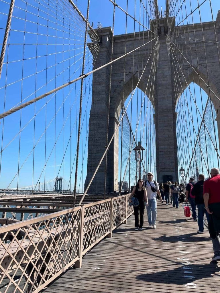 A couple walks on the Brooklyn Bridge with a blue sunny sky behind them.