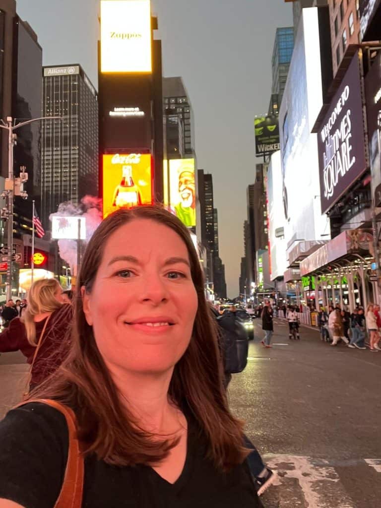 A woman smiles with Times Square brightly lit billboards and people crossing the street behind her.