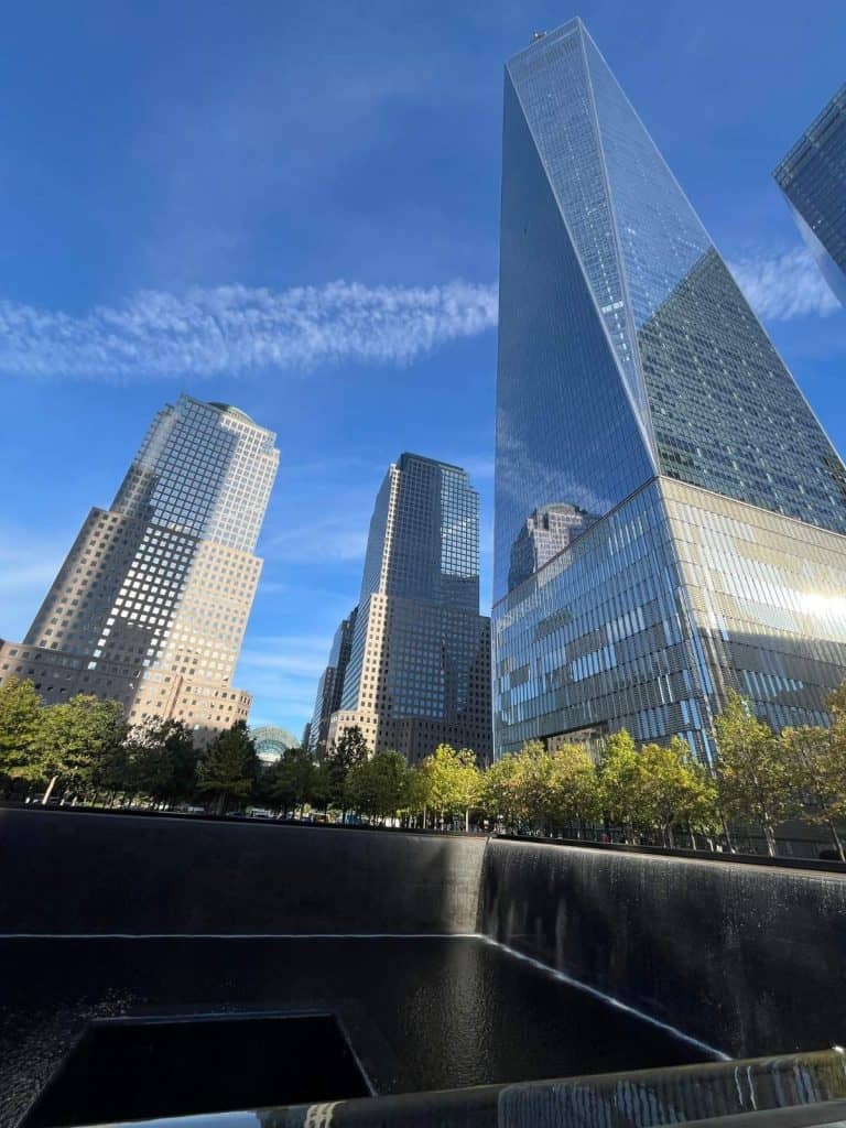 The modern 9/11 Memorial building with it's reflective exterior stands against a blue sky backdrop with the reflection pool at the base of the image.