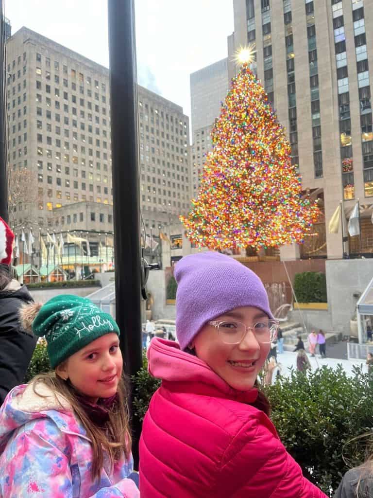 Two kids smiling while standing at the windows to the Rockefeller Center tree, which is lit up with colorful lights and has ice skating down below.