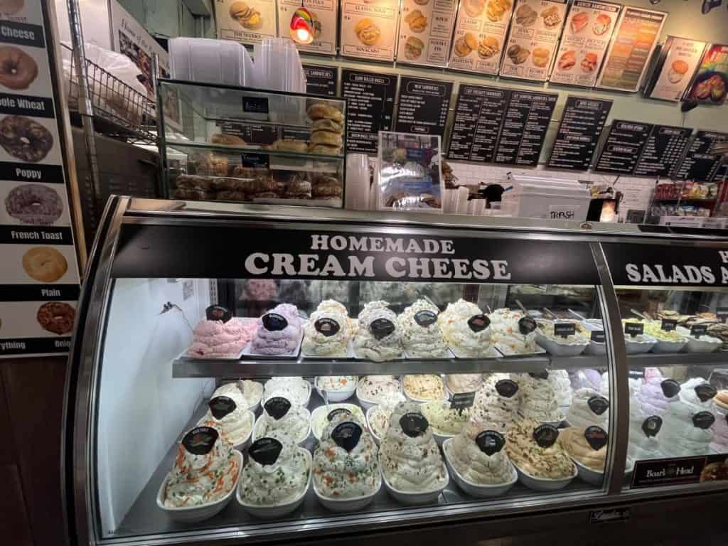 A refrigerated display case featuring rows of colorful, sculpted mounds of various homemade cream cheese flavors at a bagel store.