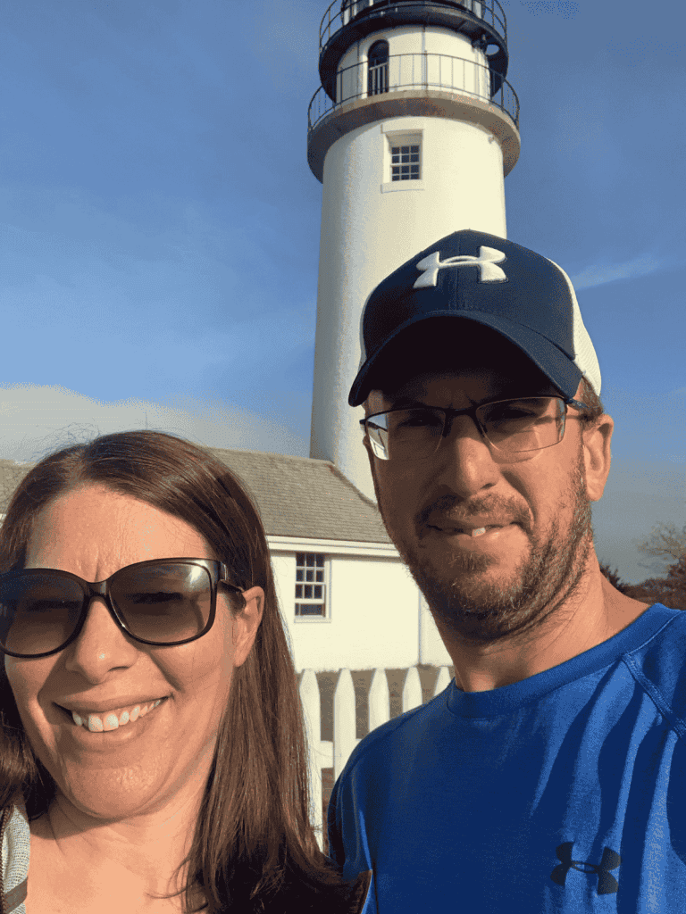 A couple standing in front of a lighthouse smiling on a blue sunny day.