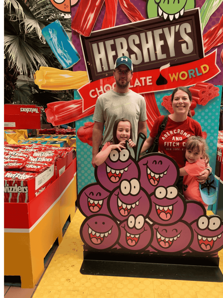 A family stands in front of a Hershey's Chocolate World display, with Twizzlers candy, and a sign with cartoon smiling grapes.